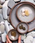 Ceramic bowls with smooth stones on a bed of larger pebbles, hand adjusting a small bowl.