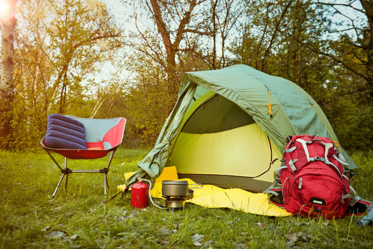 Green camping tent with a red backpack, portable stove, and a red and blue chair on grassy field.