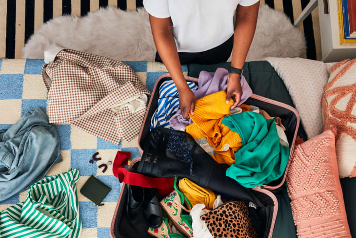 overhead shot of a messy suitcase on a bed with blue and white checkered sheets and person going through the clothes in suitcase