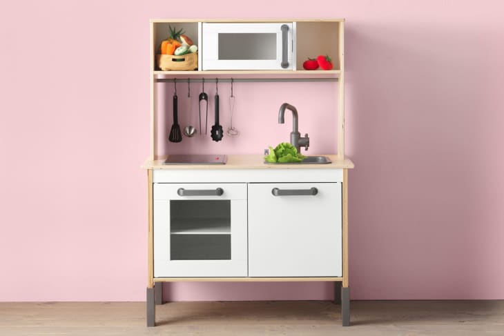 Toy kitchen set with a microwave, sink, oven, utensils, and vegetables on a pink wall background.
