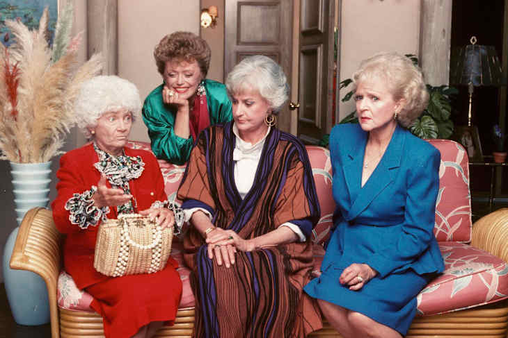 Four women sitting on a floral sofa, one in a red outfit holding a woven bag, others in colorful dresses, with pampas grass decor.