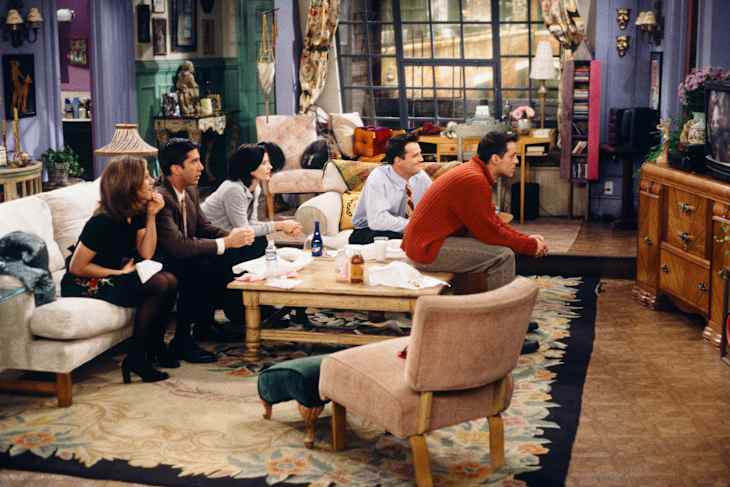 Five people sitting in a cozy living room watching TV, surrounded by eclectic decor and a patterned rug.