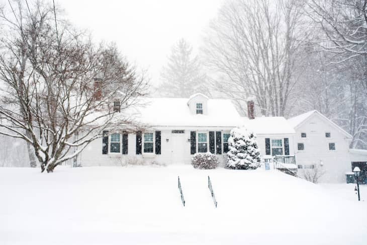 Snow-covered house with black shutters, surrounded by trees during a heavy snowfall.