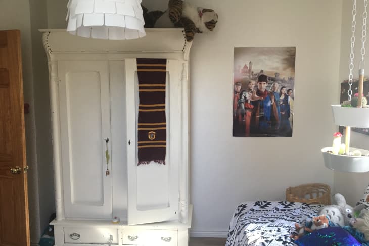 White wardrobe with a striped scarf, a tiger plush on top, a fantasy poster, and a bed with patterned bedding and stuffed animals.