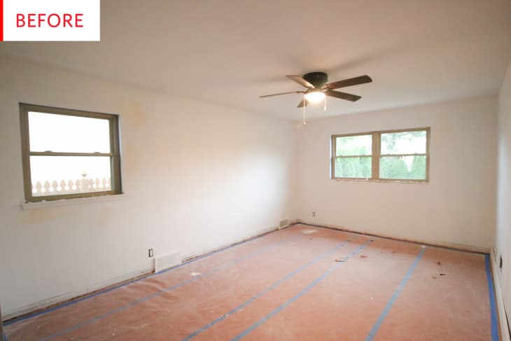 Empty room with white walls, ceiling fan, two windows, and floor covered with protective paper.