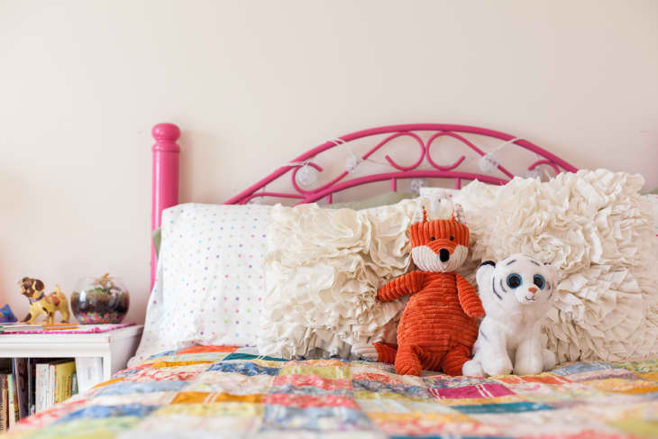 Pink metal bed with ruffled pillows, orange fox and white tiger plush toys, colorful patchwork quilt, and books on a side table.