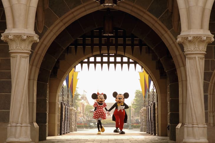 Mickey and Minnie Mouse walking hand in hand under a stone archway at a theme park entrance.