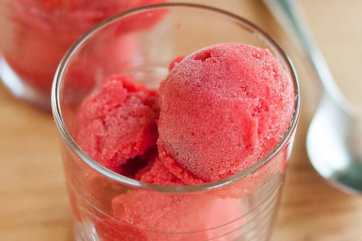 Bright red sorbet scoops in a clear glass, with a wooden surface and a spoon in the background.