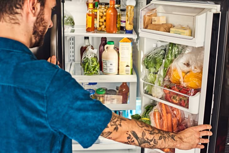 Man opening refrigerator. You can see lettuce, green onions, carrots, and other produce in the door. The rest of the fridge is full of food.
