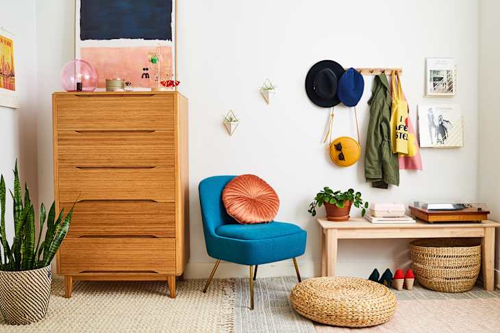 Mid-century modern room with a wooden dresser, blue chair, wall hooks with hats, and a woven basket on the floor.
