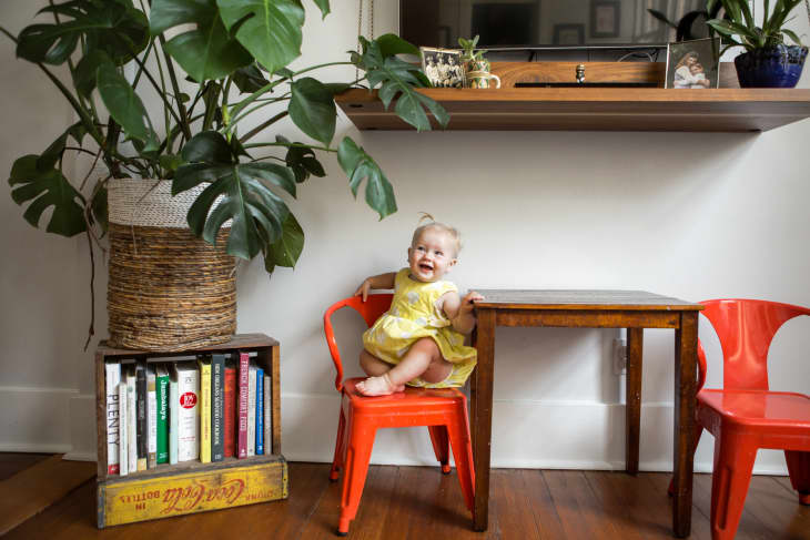 Toddler in a yellow dress sitting on a red chair next to a wooden table, with a large potted plant and books nearby.