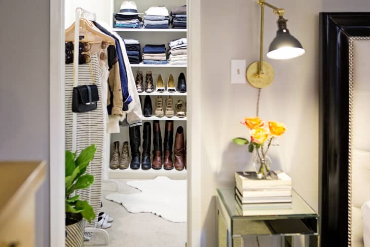 Walk-in closet with organized shelves of folded clothes, shoes, and boots; adjacent room with a lamp, books, and orange flowers.