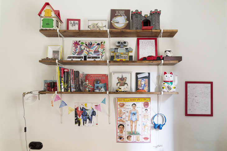 Wooden shelves with books, toys, a cat figurine, and colorful art. A body parts poster and blue headphones hang below.