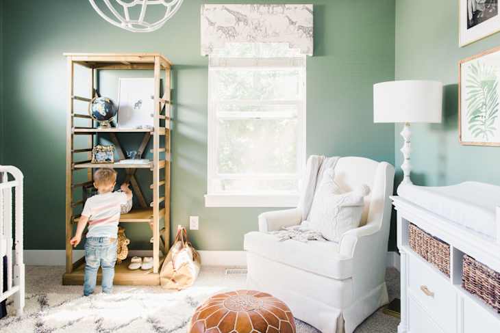 Child reaching for toys on a wooden shelf in a green nursery with a white armchair, lamp, and changing table.