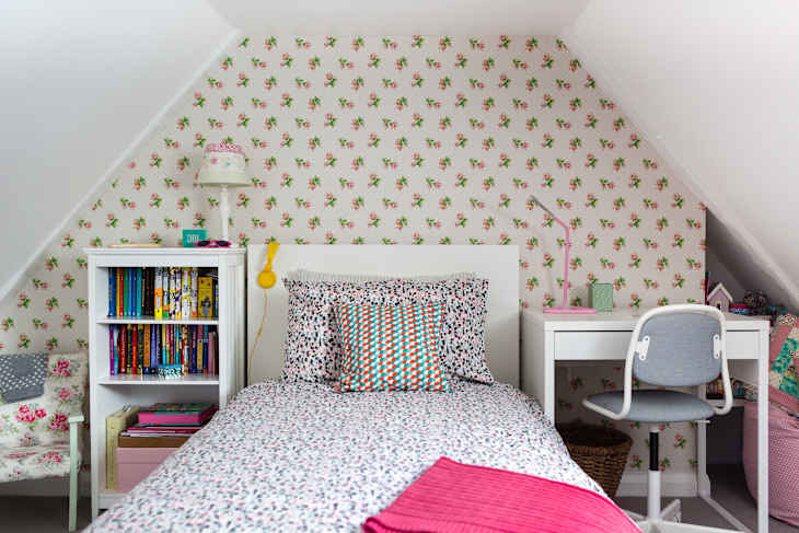 Attic bedroom with floral wallpaper, single bed, colorful pillows, bookshelf, and a white desk with a gray chair.