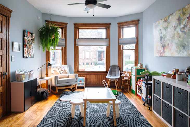 Children's playroom with a small table, chairs, toys, storage bins, and a cozy reading nook by large windows.