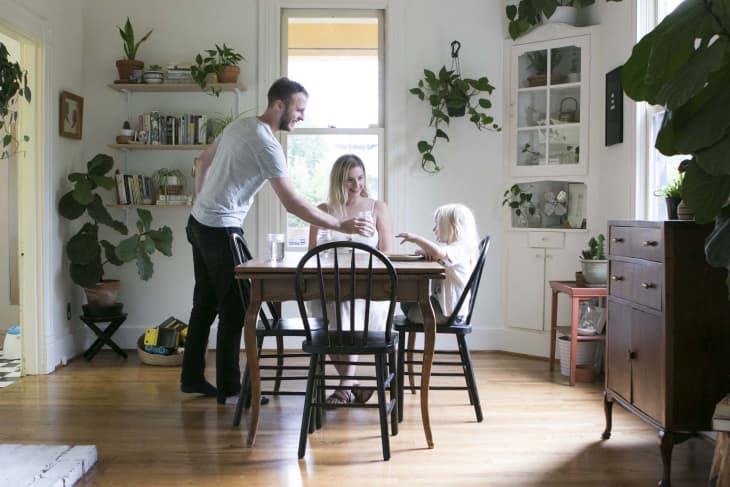 Family dining in a plant-filled room with wooden table, black chairs, and bookshelves.