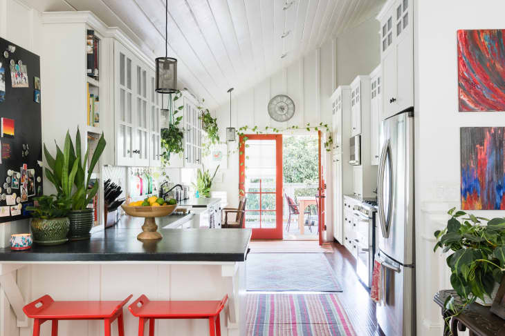 Bright kitchen with red stools, plants, fruit bowl, and glass cabinets, leading to a patio through red-framed doors.