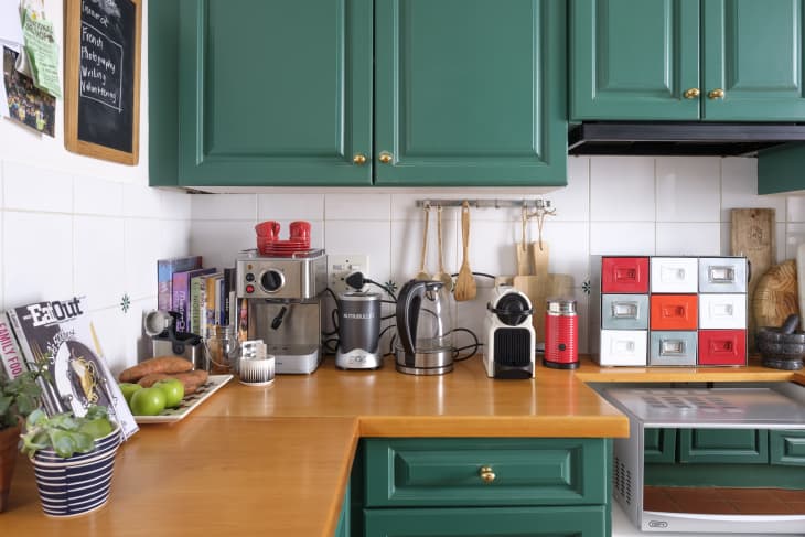 Green kitchen cabinets, wooden countertop with coffee machines, cookbooks, and colorful storage drawers.