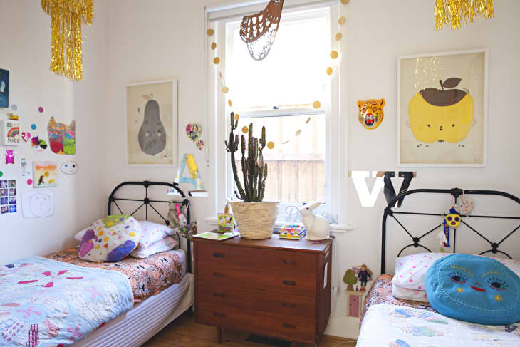 Children's bedroom with twin beds, colorful pillows, wall art, and a cactus on a wooden dresser.