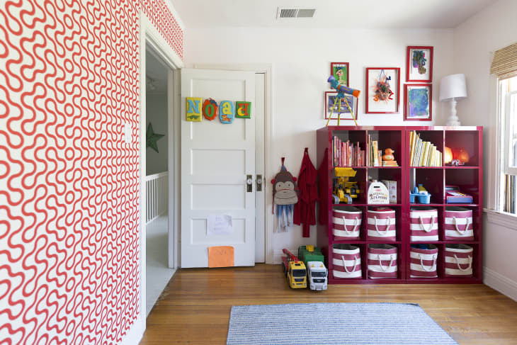 Kids' room with red patterned wallpaper, red storage shelves, toys, books, and framed art on the wall.