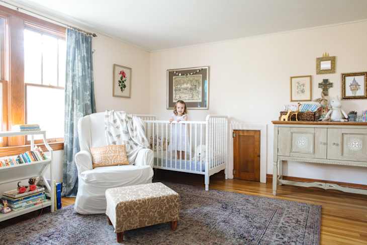 Child standing in a white crib in a nursery with a white armchair, bookshelf with toys, and a decorative sideboard.