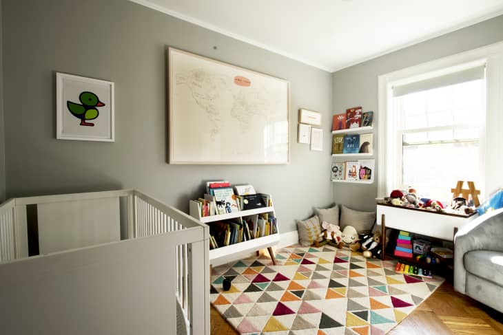 Nursery with gray walls, colorful geometric rug, crib, bookshelves, toys, and framed duck artwork.