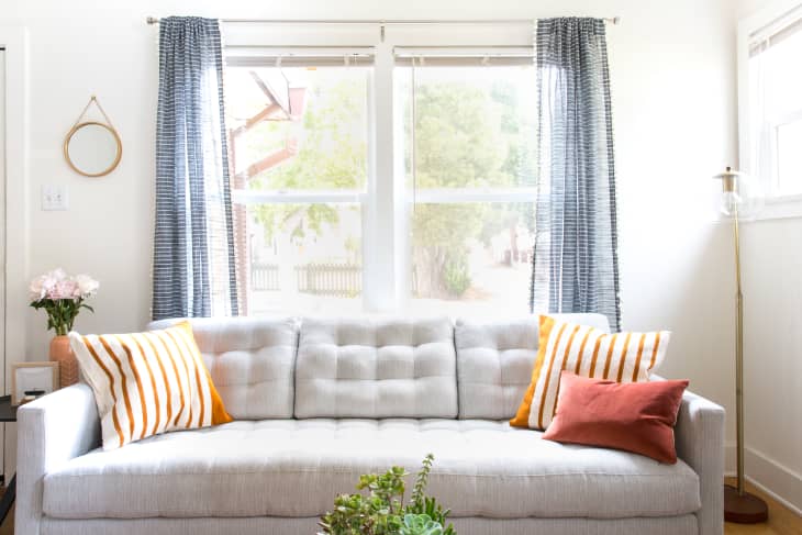 Gray tufted sofa with striped orange pillows, blue curtains, and a floor lamp in a bright living room.