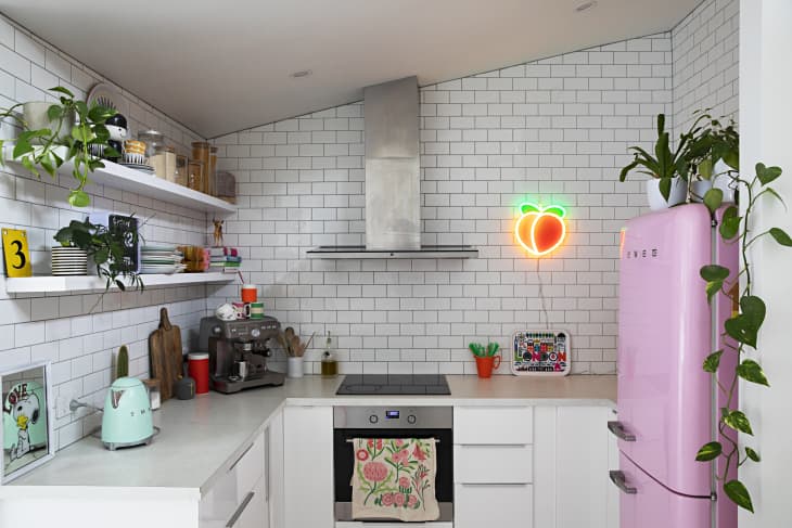 Modern kitchen with white subway tiles, pink fridge, neon peach sign, plants, and open shelves with dishes and jars.