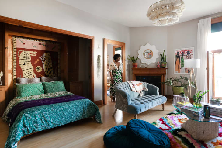 Cozy bedroom with a colorful bedspread, tapestry, blue tufted sofa, plants, and a woman in a floral dress by the door.