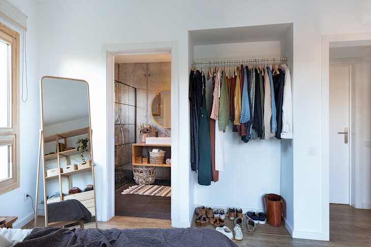 Bedroom with a floor mirror, open closet with hanging clothes, and view into a modern bathroom with a round mirror.