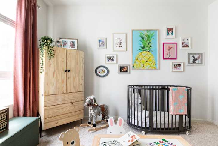 Nursery with a round black crib, colorful wall art, wooden wardrobe, rocking horse, and pink curtains.