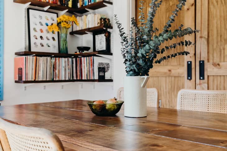 Dining table with eucalyptus in a white vase, fruit bowl, and wicker chairs, shelves with books and art in the background.
