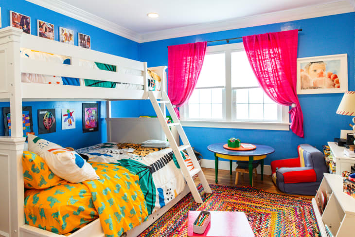 Bright children's room with blue walls, white bunk bed, colorful bedding, pink curtains, and a multicolored rug.