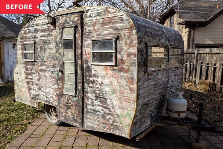 Weathered vintage camper with peeling paint, parked on a brick path beside a propane tank.