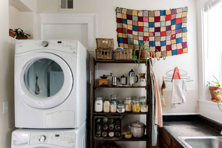 Laundry room with stacked washer and dryer, wooden shelves with jars, plants, and a colorful patchwork wall hanging.