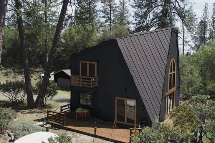 A-frame cabin with dark exterior, wooden deck, and large arched window, surrounded by trees and greenery.