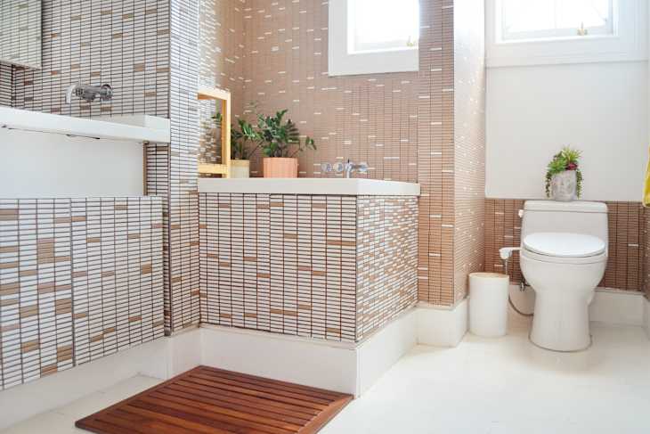 Bathroom with brown mosaic tiles, white fixtures, wooden bath mat, and potted plants on the sink and toilet.