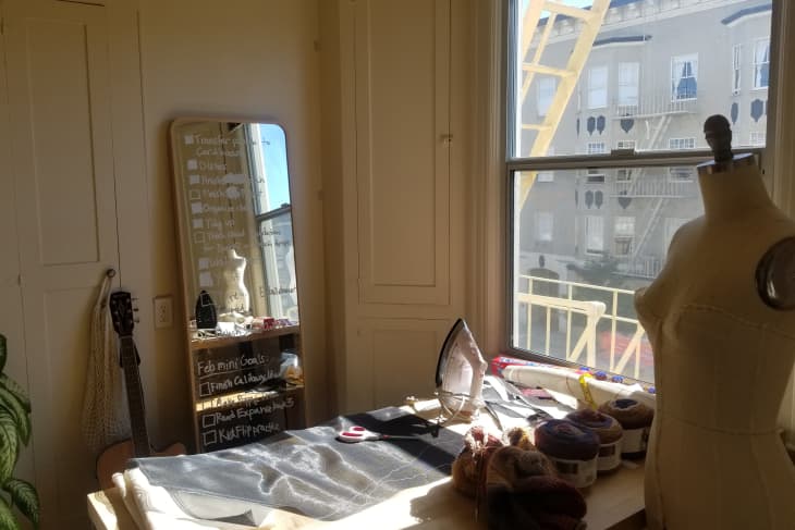 Sewing room with mannequin, fabric, yarn, iron, and a guitar by a window with a view of a building and fire escape.