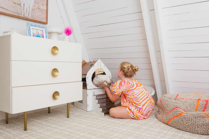 Child in colorful dress playing with a cat in a cozy attic room with a white dresser and a textured pouf.