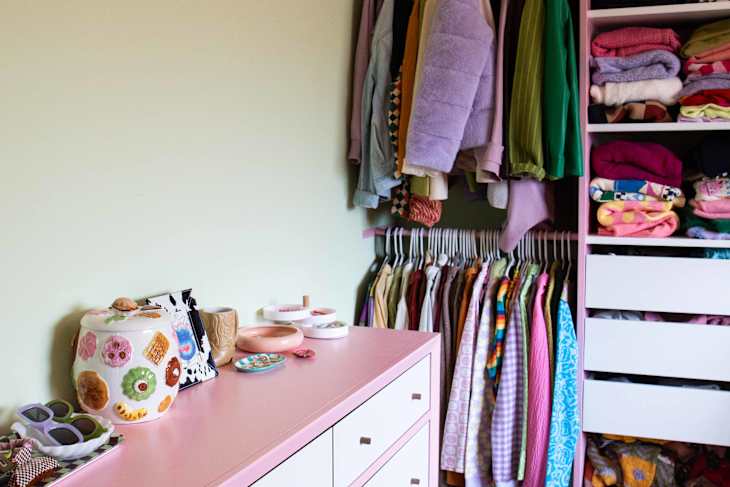 Pink dresser with decorative jars and trinkets beside a closet with colorful clothes on hangers and shelves.