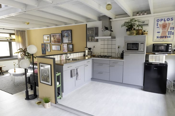 Kitchen with butcher block countertops, gray cabinets and drawers, white floor, white tile backsplash, plants, art. View into living area with pale orange wall, black and white striped area rug, gallery wall. Ceiling is white with exposed white beams