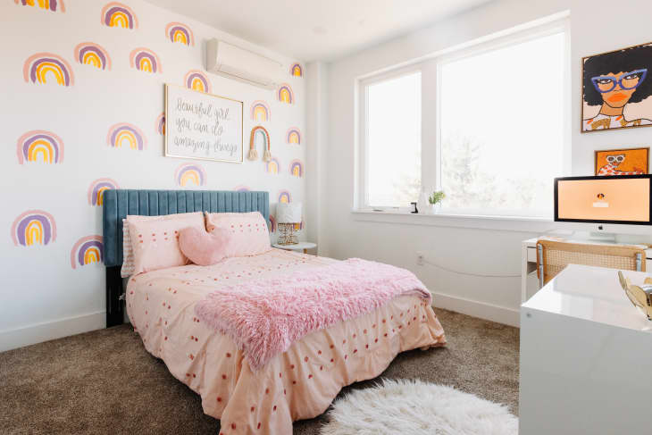 Bedroom with rainbow wallpaper, blue headboard, pink bedding, fluffy rug, and a desk with a computer and artwork.