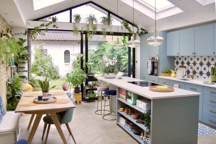 Bright kitchen with blue cabinets, island, hanging plants, and large windows overlooking a garden.