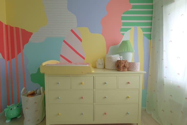 multi colored splatter wall mural with stripes throughout, yellow dresser, yellow changing table on top, toys, basket of toys, light airy white curtains, green pleated lamp shade, wood floors