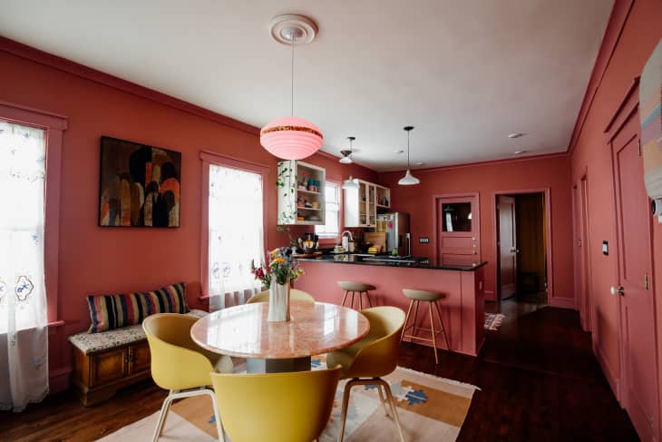 Pink pendant light over pink marble table in dining room surrounded by yellow dining chairs in warmed toned dining and kitchen area.