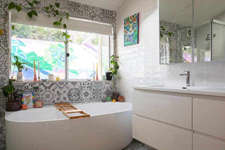 glossy white subway tile, white tub, plant on windowsill, grey and white unique designed tile, candlesticks, rubber ducky, small pots, grey and white patterned rug matching tile, white two drawer vanity, hanging plants, angled ceiling, wood tub shelf, mirror medicine cabinet