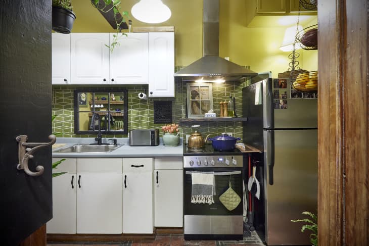 Kitchen with pale yellow green walls, green tile backsplash, white cabinets with black pulls, hanging plants, pale gray countertops, worn tile floor, lots of antique and decorative accents