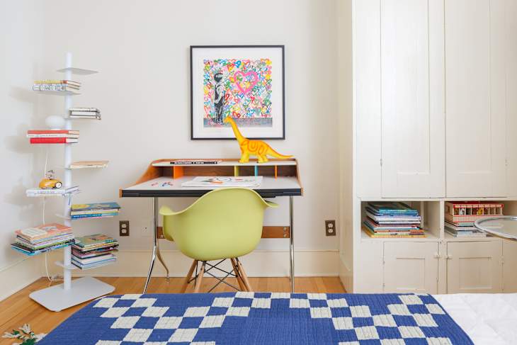 Art print mounted above a desk in a child's bedroom with green armchair under desk and toy giraffe set  on top of desk. Books stacked on bookshelf in corner.