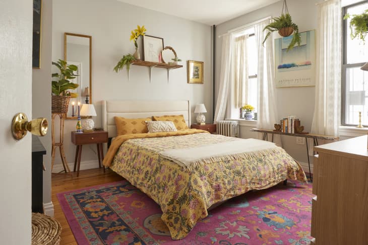Plant filled bedroom with neatly made bed topped with pale yellow quilt. Colorful pink rug lines the floor.